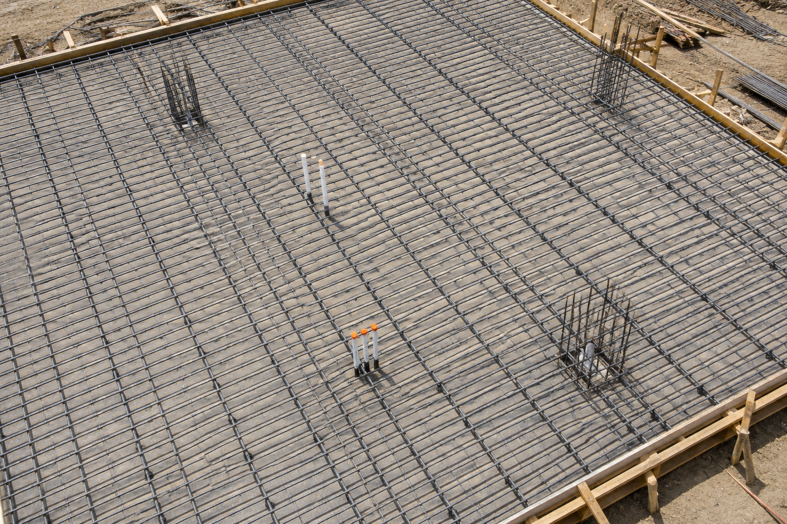Steel rebar grid installed across a concrete slab foundation with utility pipes and vertical reinforcement bars